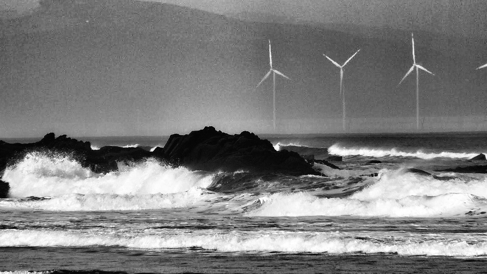 Fotografía en blanco y negro de un paisaje costero tormentoso.  Olas blancas rompen contra unas rocas oscuras en primer plano. Al fondo, se aprecian tres aerogeneradores marinos en el horizonte, bajo un cielo gris. La imagen transmite una sensación de poderío de la naturaleza y la integración de la energía renovable en el entorno.