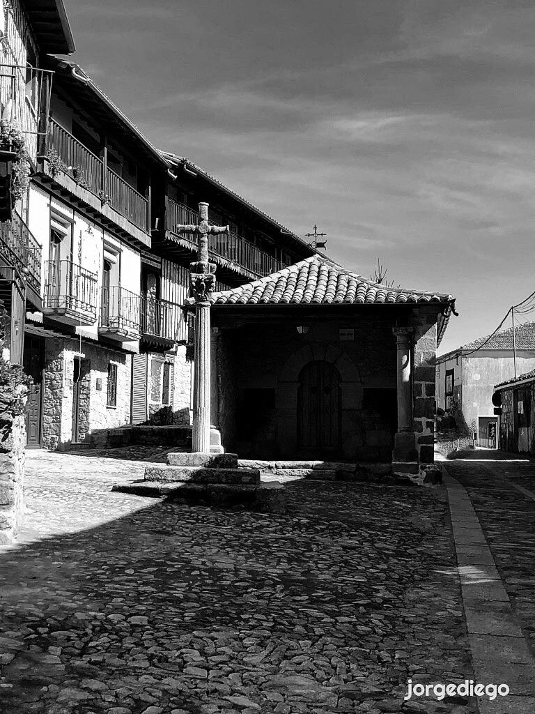 Fotografía en blanco y negro de una plaza adoquinada en Mogarraz, Salamanca. En el centro de la plaza se encuentra una pequeña capilla o estructura con techo de teja y una cruz de piedra en un pilar alto frente a ella. Al fondo, un edificio de varios pisos con balcones de hierro forjado se extiende a lo largo del lado izquierdo de la imagen. La luz del sol crea fuertes contrastes de sombra y luz en la escena. El ambiente es tranquilo y evoca la arquitectura tradicional de un pueblo histórico.