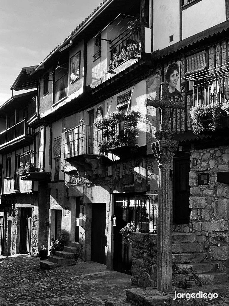 La imagen muestra una calle empedrada de Mogarraz, Salamanca, con edificios de piedra de varios pisos con balcones de hierro forjado.  Las fachadas de las casas están adornadas con retratos en blanco y negro de personas, que parecen estar integrados en la arquitectura. Una cruz de piedra destaca en primer plano, junto a una de las casas. La escena está bañada por la luz solar, creando sombras y contrastes que acentúan la textura de la piedra y la arquitectura del lugar.  El ambiente es tranquilo y evoca un pasado histórico.