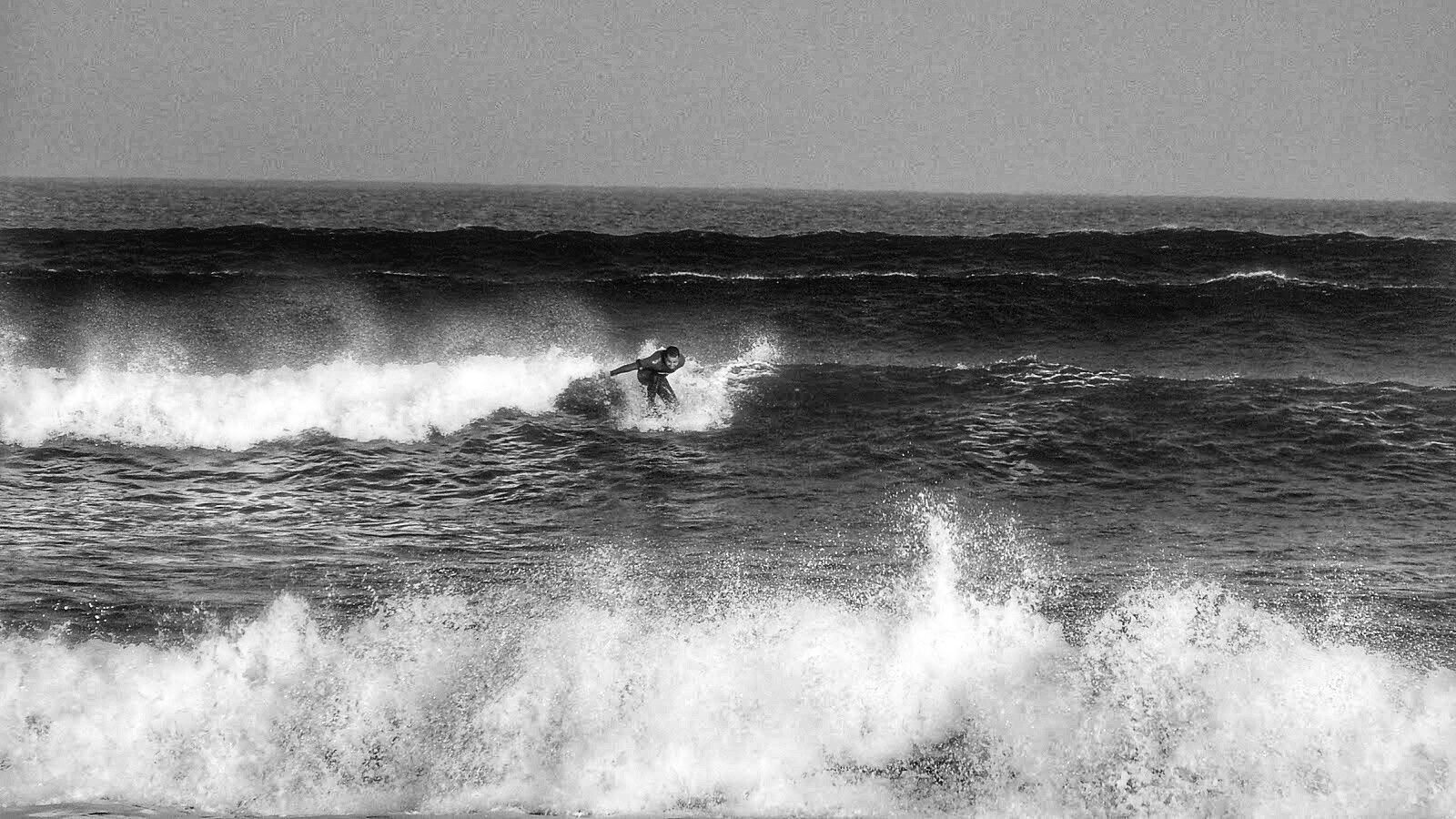 Foto en blanco y negro de un surfista surfeando una ola en la playa de Sopelana, Euskadi. El surfista está de perfil, ligeramente inclinado hacia adelante, sobre una tabla de surf. Las olas son moderadamente grandes y rompientes, con espuma blanca visible. El océano es oscuro y el cielo está despejado. La imagen tiene un tono monocromático que le da una atmósfera tranquila y dramática.