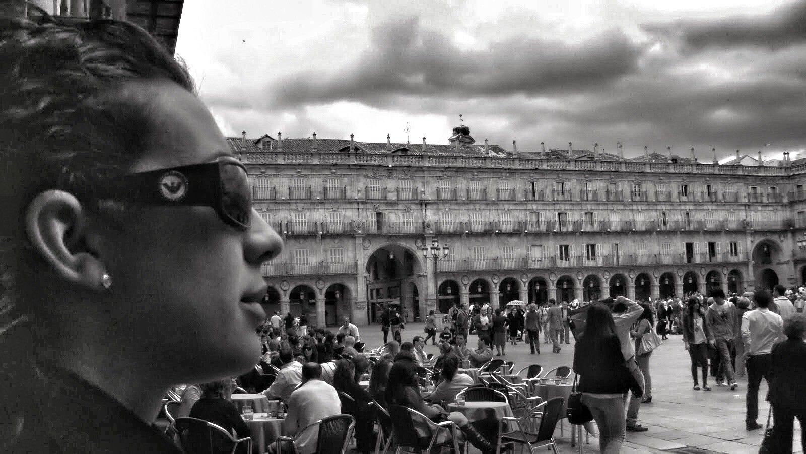 Una foto en blanco y negro de una mujer joven, vista de perfil, que lleva gafas de sol y mira hacia la Plaza Mayor de Salamanca. La plaza está llena de gente, y el cielo está nublado. La mujer está en primer plano, y el edificio y la gente están en segundo plano.