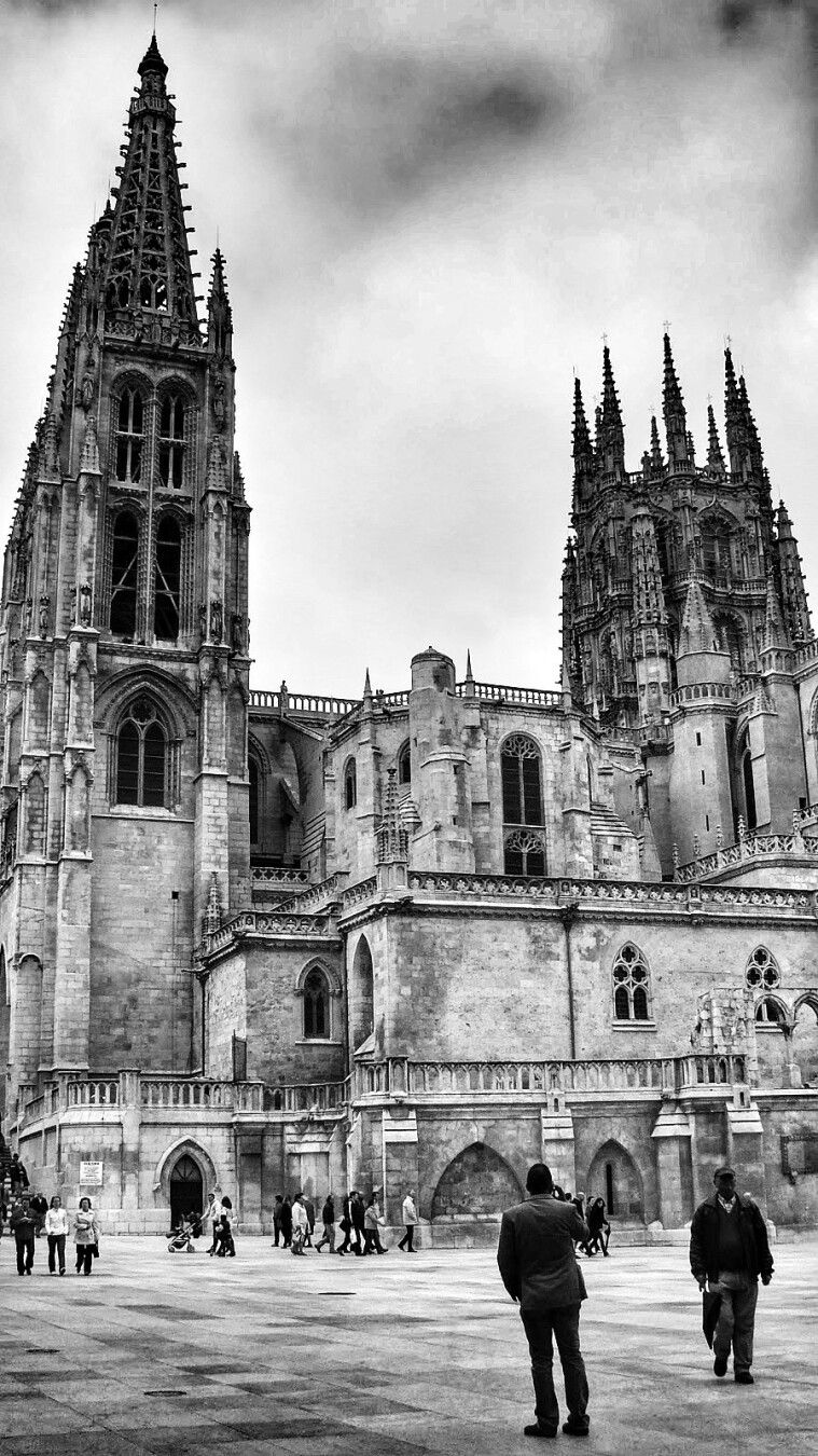 Fotografía en blanco y negro de la Catedral de Burgos, España.  Se muestra una vista de la parte trasera de la catedral, con sus imponentes torres góticas, una alta y puntiaguda, la otra más baja y con múltiples pináculos.  La fachada principal de la catedral es de piedra, con intrincados detalles arquitectónicos y arcos apuntados.  En la plaza frente a la catedral hay varias personas caminando, creando una sensación de escala. Dos hombres están de espaldas a la cámara, cerca del primer plano. El cielo está nublado.