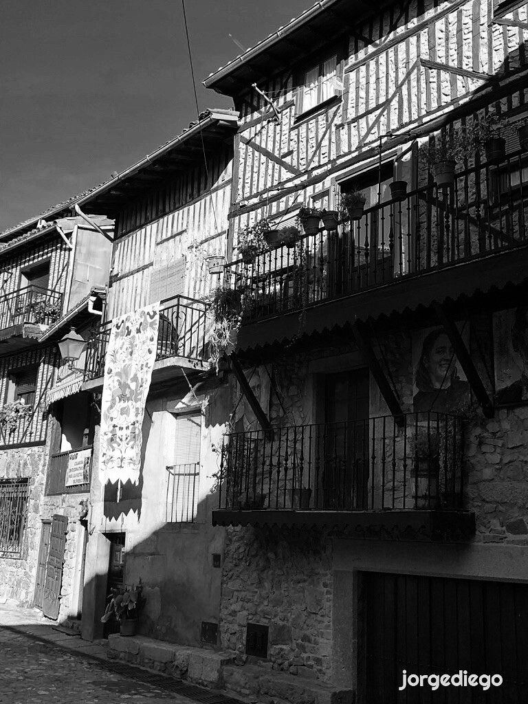 Fotografía en blanco y negro de una fila de casas antiguas de piedra en Mogarraz, Salamanca, con entramados de madera visibles, se alinean en una calle estrecha. Las casas tienen balcones con barandillas de hierro forjado, adornados con macetas. Una tela o tapiz está colgado en el exterior de una de las casas. La escena está iluminada por una luz solar lateral, creando fuertes contrastes de luz y sombra. Se aprecia un ambiente rural y tranquilo.