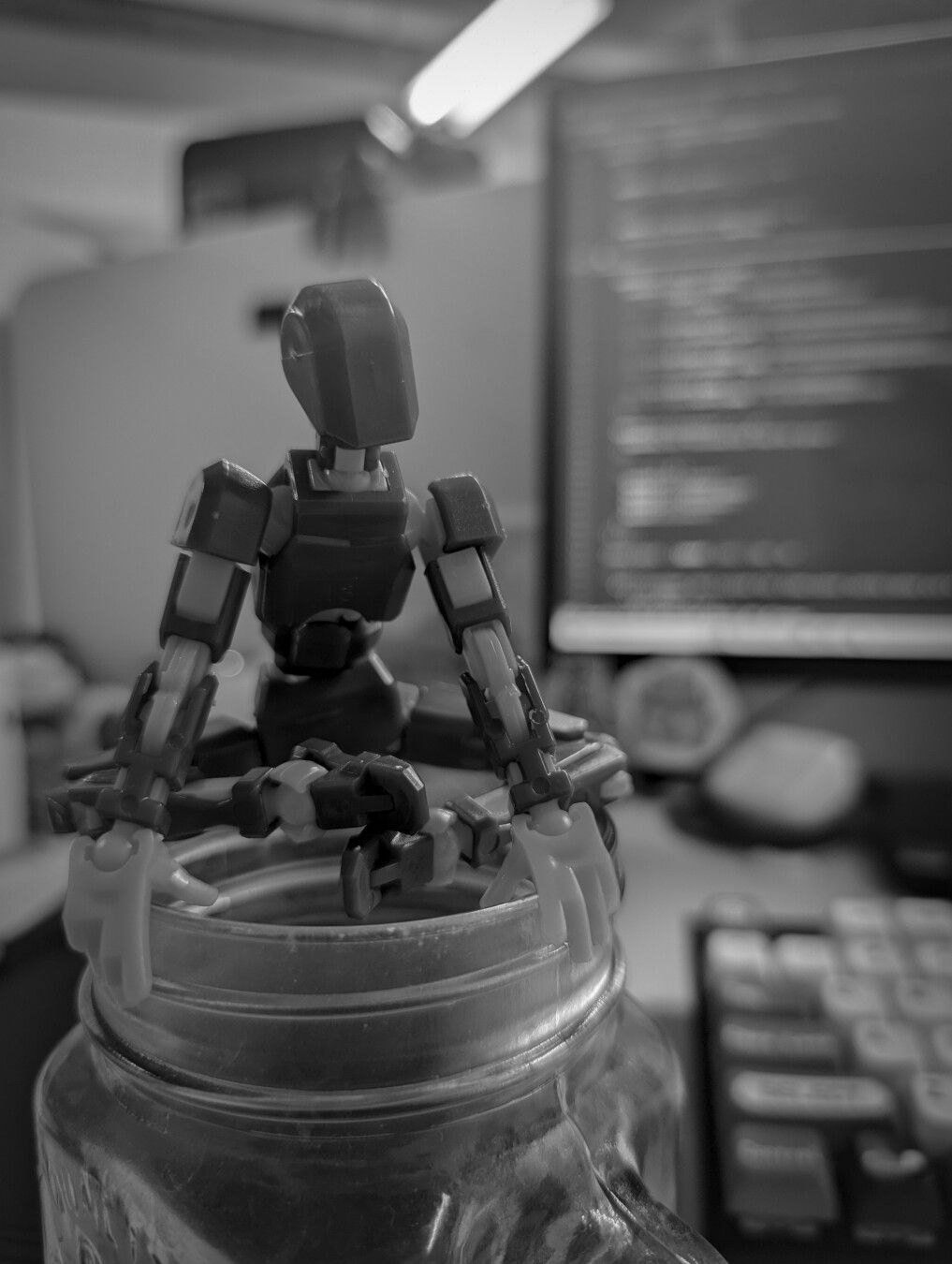 Figura de acción de un robot en posición de meditación sobre un frasco de vidrio, con un monitor de computadora de fondo.