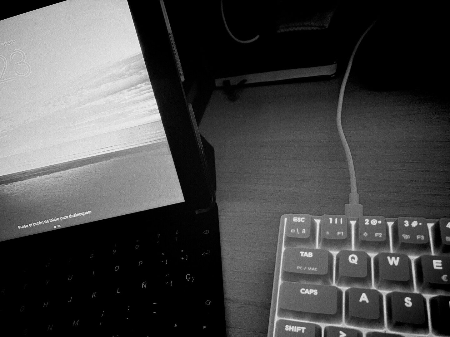 En blanco y negro. Se ve un fragmento de teclado mecánico y un fragmento de una tablet abierta con su teclado y la pantalla encendida.