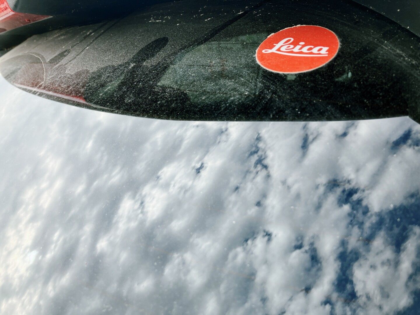 Cristal trasero de un coche con el reflejo de las nubes y una pegatina roja circular de Leica.