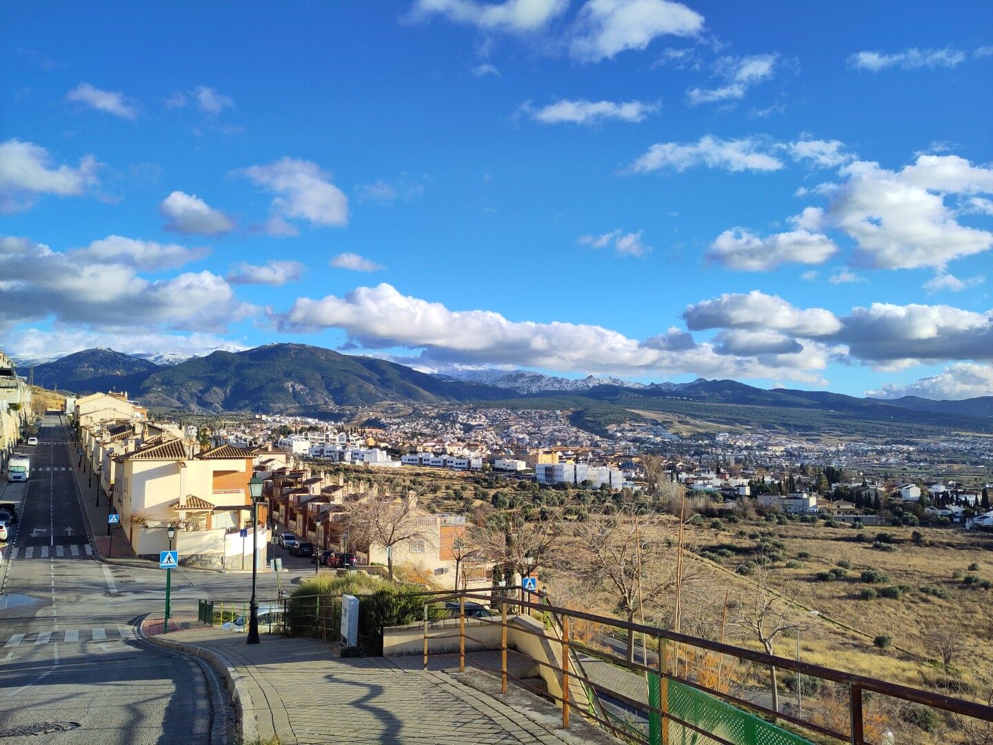 Fotografía donde en el centro se ve una calle bajando y a la derecha una barandilla, con vistas a los pueblos del sureste de Granada. De fondo se ve Sierra Nevada blanca, aunque parcialmente tapada por nubes. Hay un cielo azul tapado con nubes voluminosas y con muchas sombras