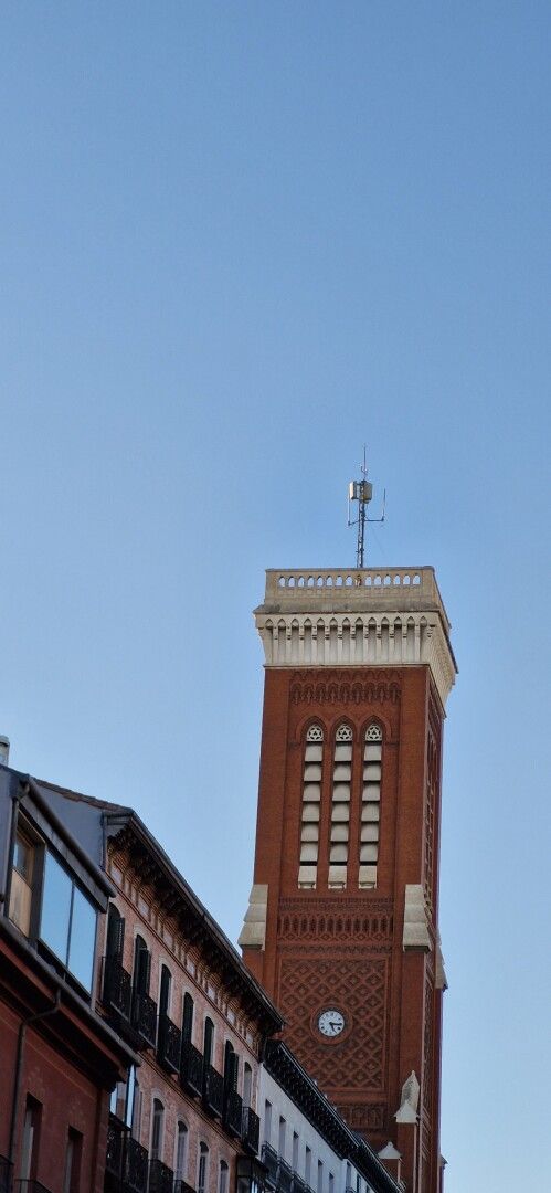 Fotografía donde se ve una torre con un reloj y la fachada y el tejado de un edificio. El cielo está despejado y azul