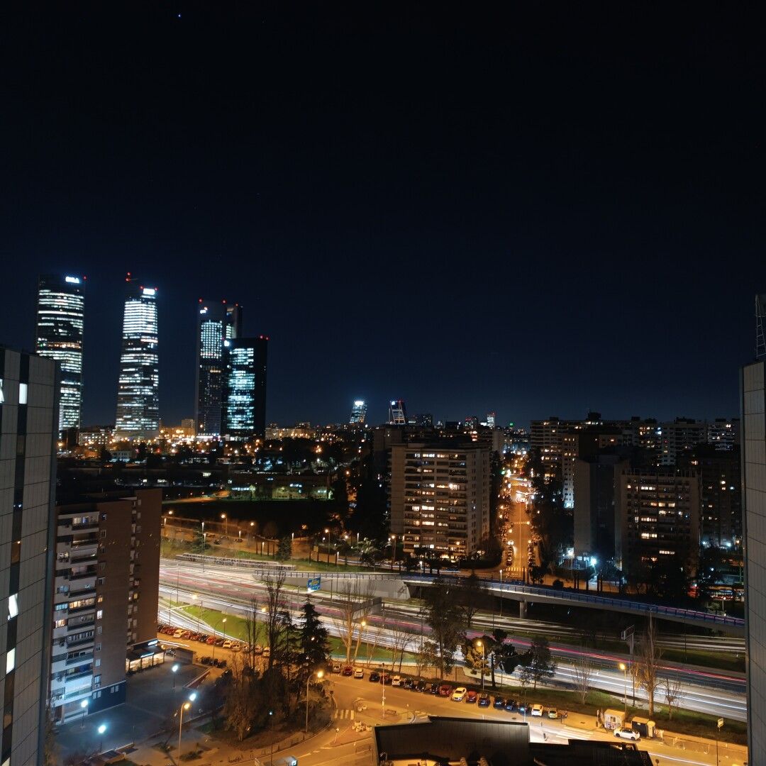 Fotografía nocturna desde una ventana en la que se ven edificios con luces, una carretera y el cielo oscuro. se identifican varias de las torres de la Castellana en Madrid