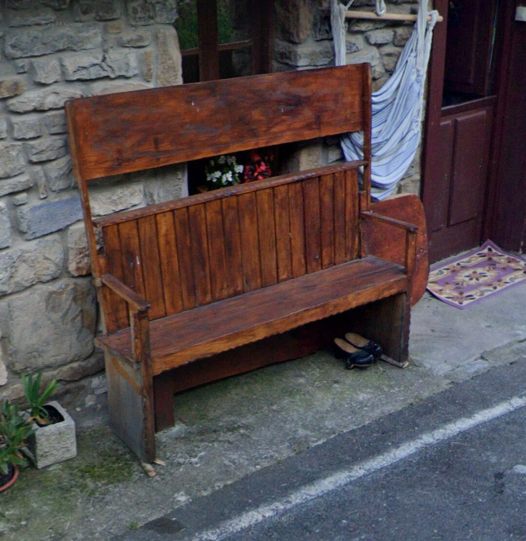 Esto se llama ¨escaño¨, era muy habitual en las casas asturianas, Y se situaba en la cocina cerca del llar. Es un banco de madera para 2, 3, 4 personas. Tiene una mesa abatible que normalmente esta levantada, quedando por encima de la cabeza de los que están sentados en él. A la hora de comer se sentaban y se bajaba la mesa abatible. Con la mesa bajada sólo se puede entrar o salir agachándose por debajo de la mesa. Como bien supondréis, eran los paisanos y los abuelos los que comían ahí, mientras que la madre era la que servía la comida.
Éste está situado en la parte de afuera de un bar de pueblo con las paredes de piedra vista, también hay una ventana de madera, la puerta de entrada al bar, también de madera, y una tela colgada al lado de la puerta.
Como curiosidad, yo tuve alquilada una casa en ese pueblo en los ochenta, y juraría que es el mismo escaño que tenía la casa.