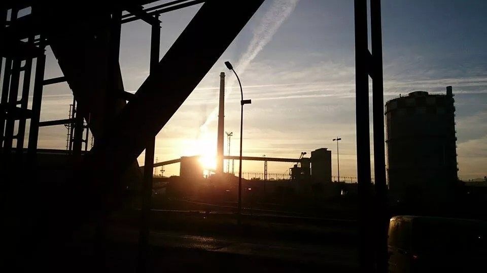 Foto en claroscuro en el que se ve el sol saliendo en la lejanía tras un edificio industrial y von una alta chimenea a su derecha. También se ven otras estructuras industriales, como un par de cintas transportadoras en altura, que convergen con el edificio tras el que sale el sol, así como una de ellas va hacia otro edificio más alto situado más a la derecha. A la derecha de dicho edificio y mucho más cerca se ven unas estructuras de soporte de otra cinta, y a su derecha y más alejado, vemos un gasómetro.
Lo poco que se ve del cielo va tomando un color azul, aunque se ven unas nubes bajas y varias estelas de aviones cruzándose. en primerísimo plano se ven dos grandes vigas de soporte cruzándose en forma de equis. lo más cercano está todo negro, pues la foto fue tomada al aire libre, aunque bajo una gran estructura que soporta varias cintas transportadoras.