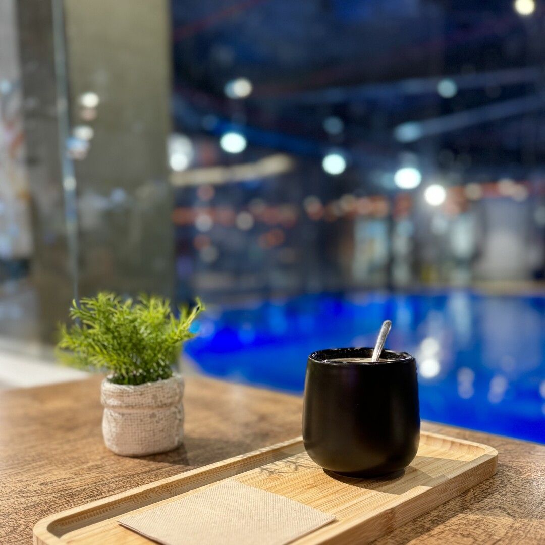 Bandejita de madera con una taza sin asa negra chulísima y una servilleta. Sobre la mesa, también de madera, hay una pequeña plantita decorativa. De fondo está el azul correcto de la piscina de buceo. 

#PepPerCoffee