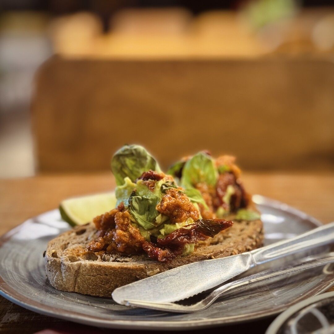 Detalle de LA TOSTADA de aguacate con tomatitos secos y pesto rojo. Está sobre pan de semillas y viene con un trocito de lima para que le eches por encima y vayas al infierno directamente porque esto tiene que se pecado.