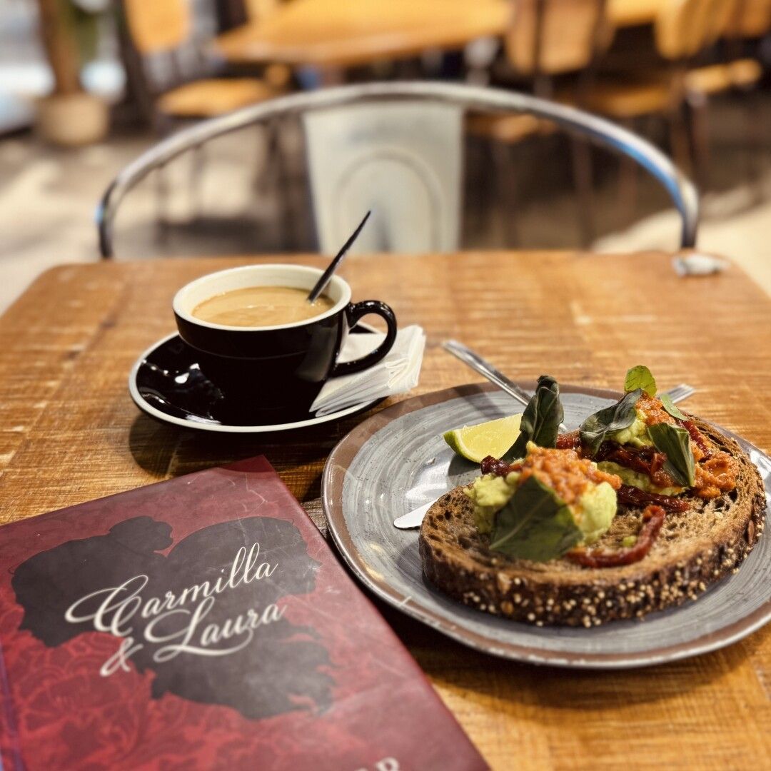 Bodegón de desayuno con libro, café súper rico con leche fría de avena en taza negra y tostada de aguacate y pesto rojo.