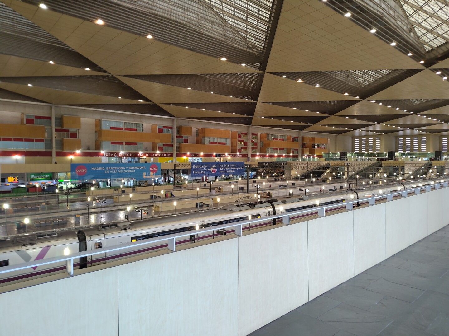 Vista desde el interior de la estación Delicias de Zaragoza desde un lateral en alto después de subir del andén de llegada. Se ven andenes con algún tren, anuncios y el techo dividido en triángulos.