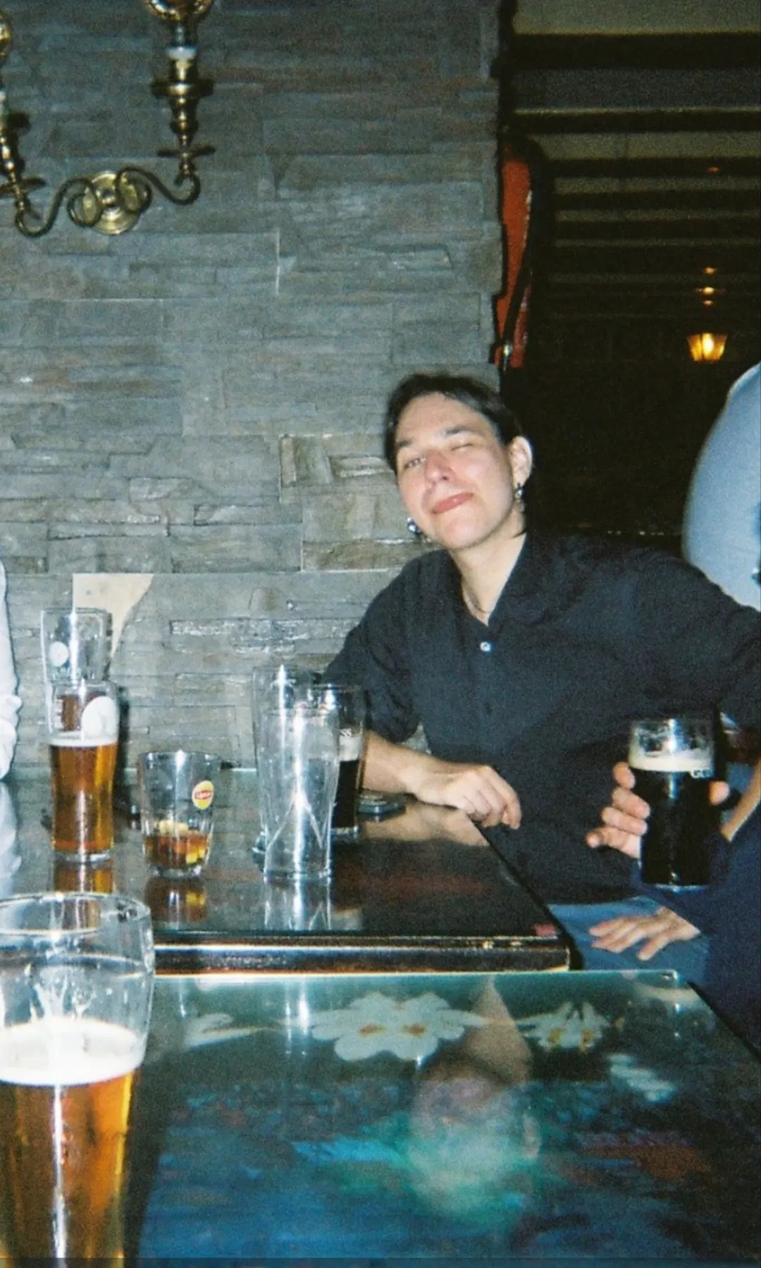 Foto del proyecto de tía que les habla. Estoy en un bar mal iluminado a última hora de la boda, sentada y con el brazo apoyado en la mesa. Llevo camisa negra y pendientes plateados. Guiño el ojo y saco la lengua.
En la mesa, cervezas varias (estábamos esperando a alguien creo recordar)