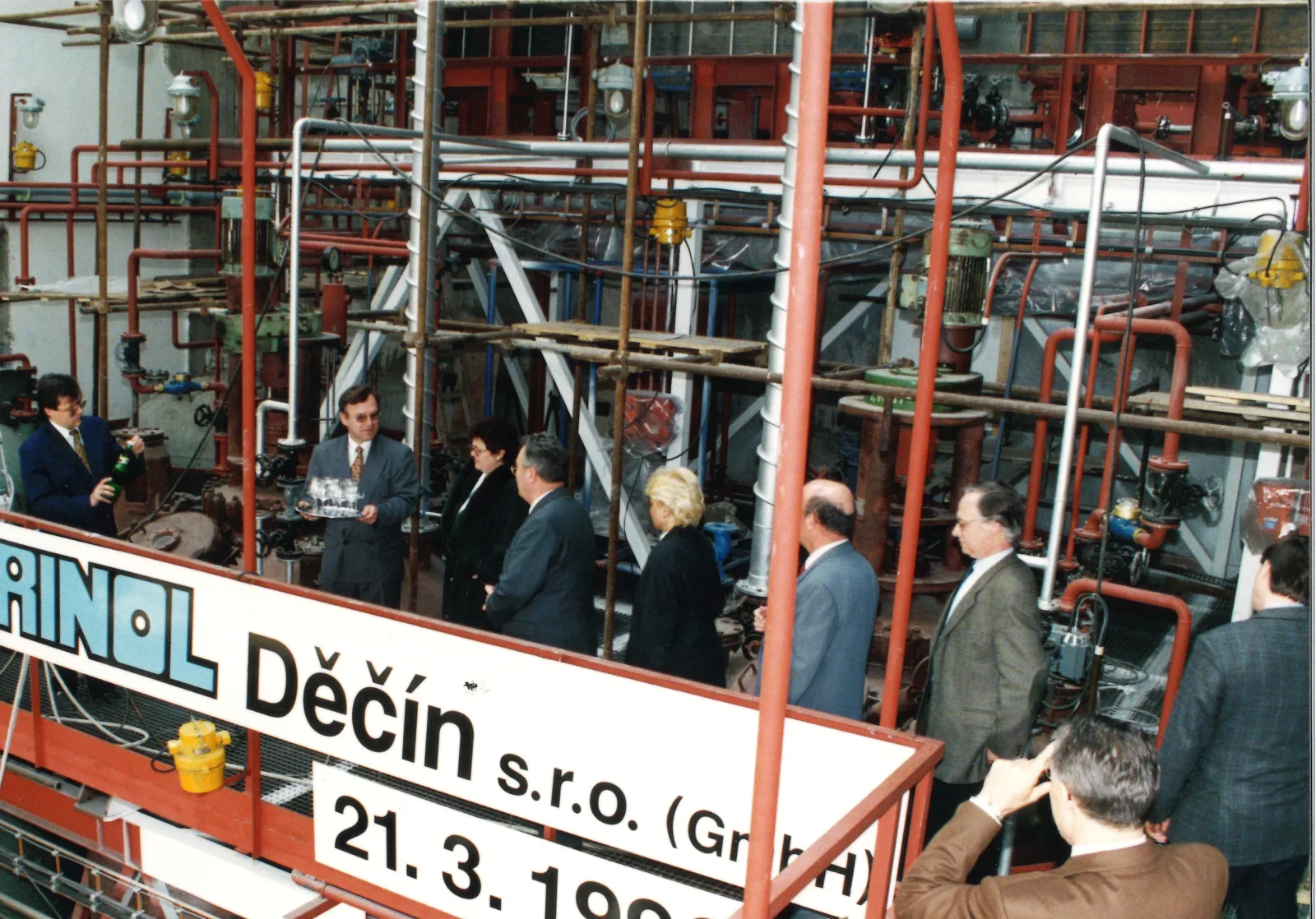 Ein Mann schenkt Sekt ein, während eine Gruppe zwischen Rohren und Maschinen in der Fabrikhalle von HABRINOL in Děčín steht.